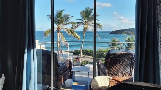 Ein Balkon mit Korbstühlen und einem Tisch, mit Blick auf einen Strand mit Palmen und dem Meer.