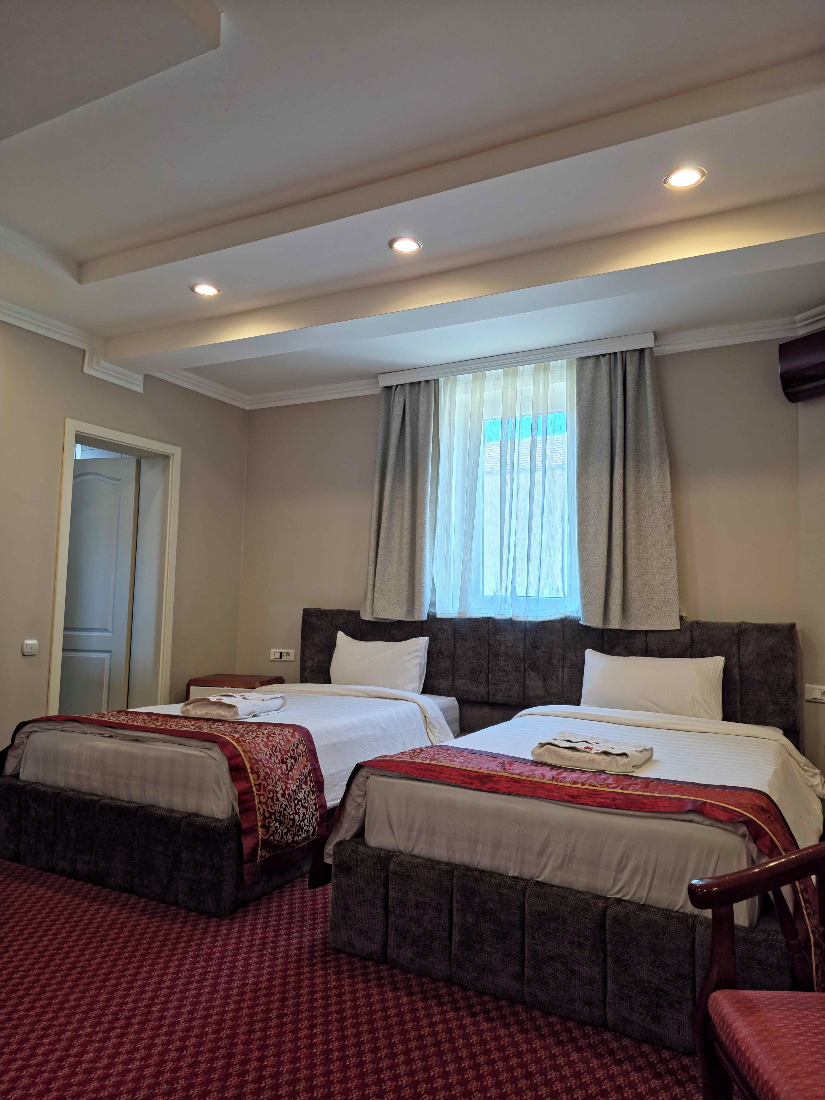 A hotel room with two beds, a large window with curtains, a door, and a carpeted floor.
