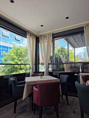 A restaurant with a table set for two, large windows with curtains, and a view of greenery and buildings outside. - Hotel Royal (Pristina)