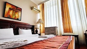 Une chambre d’hôtel avec un grand lit, un tableau au mur, un appareil de climatisation et une vue sur l’extérieur à travers de fines rideaux.