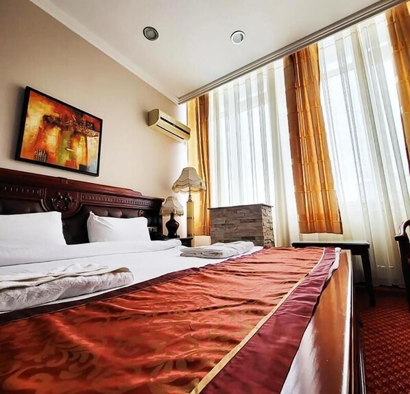 A hotel room with a large bed, a painting on the wall, an air conditioning unit, and a view of the outdoors through sheer curtains. - Hotel Royal (Pristina)