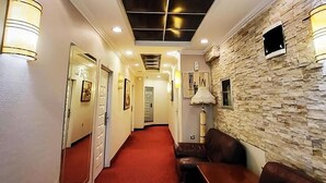 Un couloir d’hôtel avec un tapis rouge, un plafond doté d’un éclairage encastré, un mur en pierre orné d’un téléviseur et un coin salon avec des canapés en cuir.