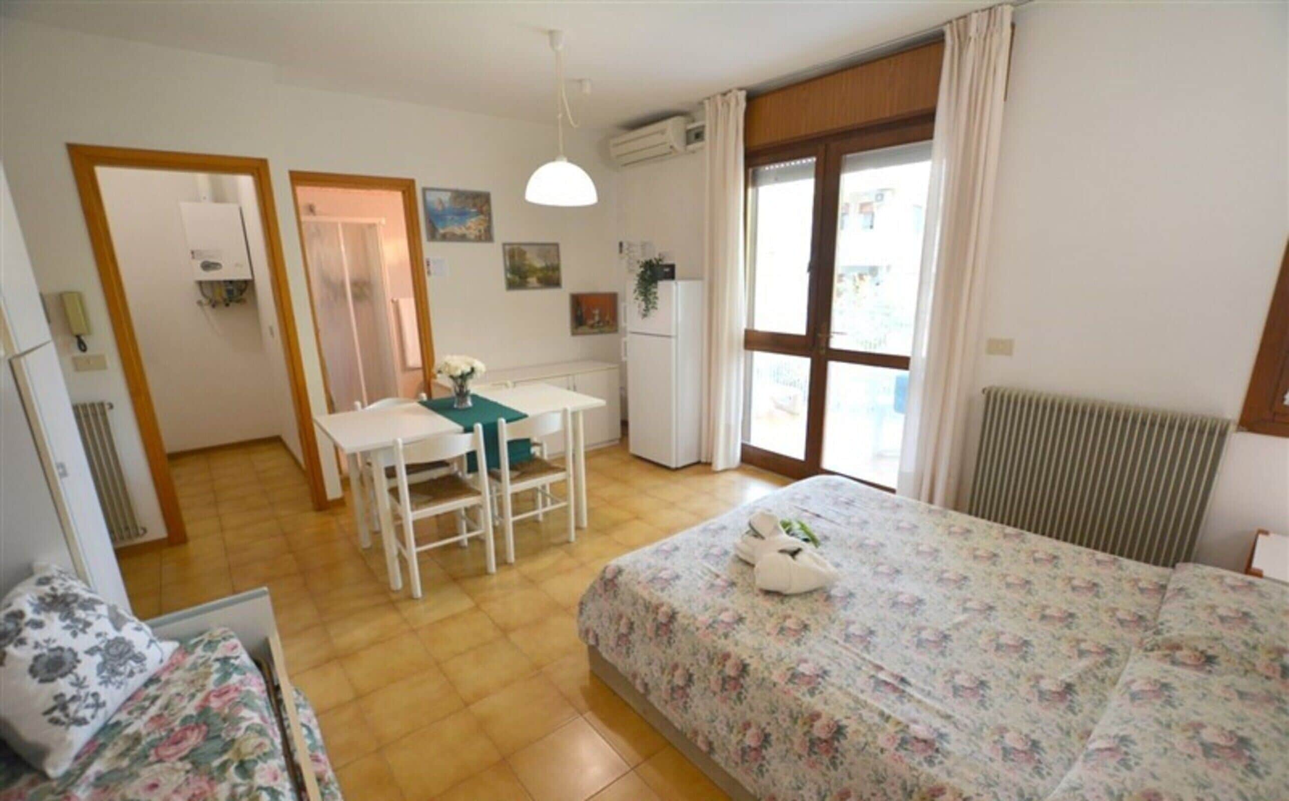 Habitación pequeña con cama, un escritorio y una silla. Se observa un radiador, una ventana con cortinas y una puerta que da a otra habitación.
