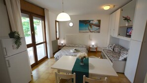 Habitación compacta con cama, una mesa con sillas, sofá, kitchenette pequeño y un ventanal grande con cortinas.