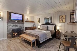 A bedroom with a stone fireplace, a bed with a headboard, a bench, a table with chairs, and a television mounted above the fireplace. - Valentine's Cabin w/WiFi • Scenic Views • Walk to Downtown • Fireplace (Bryson City)