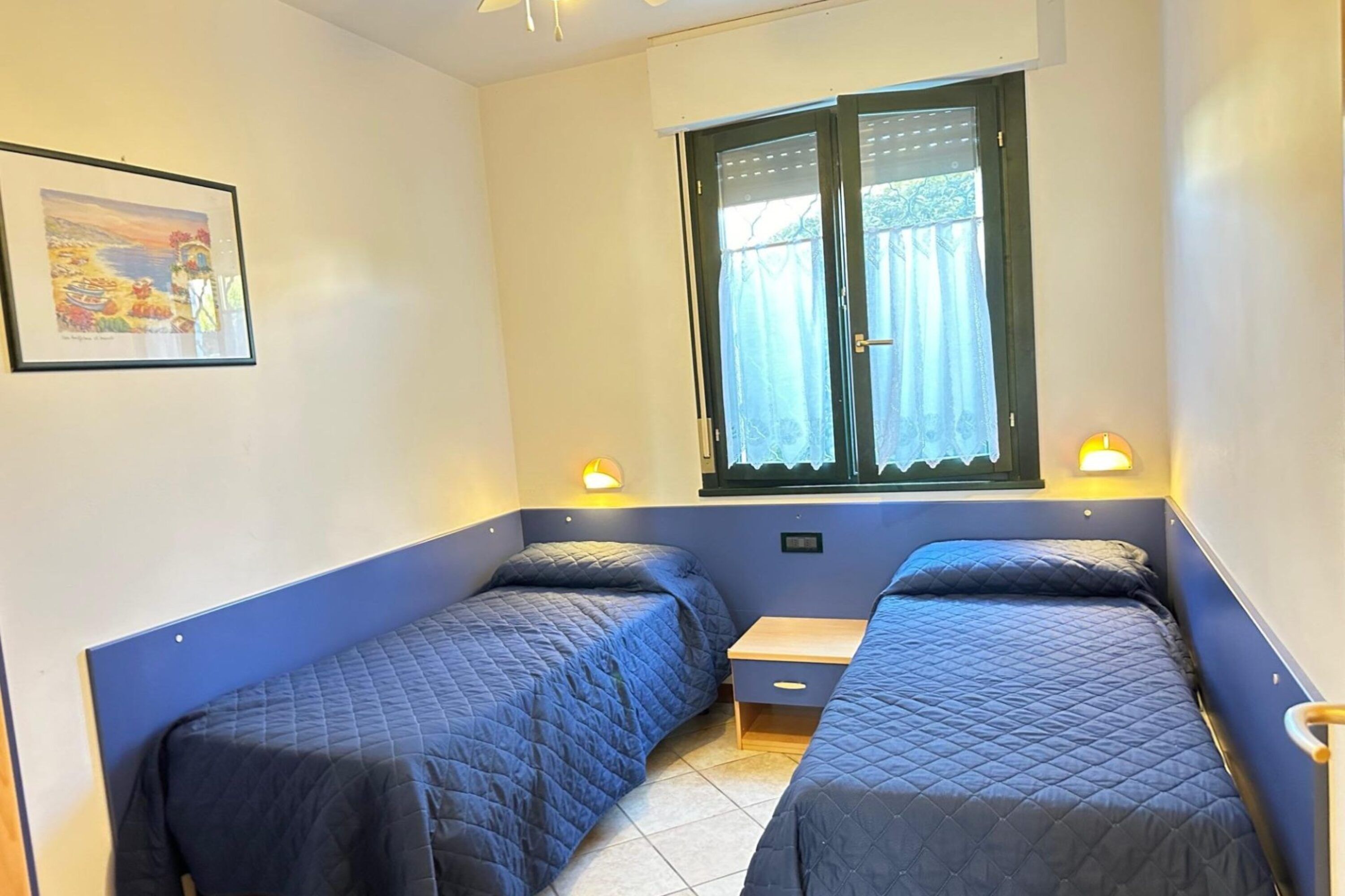 A room with two single beds, a small wooden nightstand, a window with curtains, and a wall-mounted light fixture.