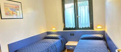 A room with two single beds, a small wooden nightstand, a window with curtains, and a wall-mounted light fixture.