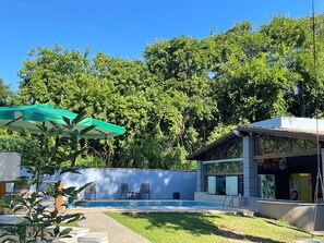 A pool area with a covered seating area, a building with a glass facade, and lush greenery in the background. - Pousada Píer 22 (Caraguatatuba)