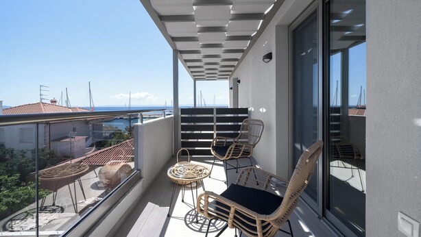Ein Balkon mit Korbmöbeln, einer Glasreling und Blick auf das Meer und Gebäude.