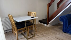 Un'area pranzo modesta con un tavolo bianco e due sedie, un telecomando sul tavolo, un divano blu e una scala in legno.