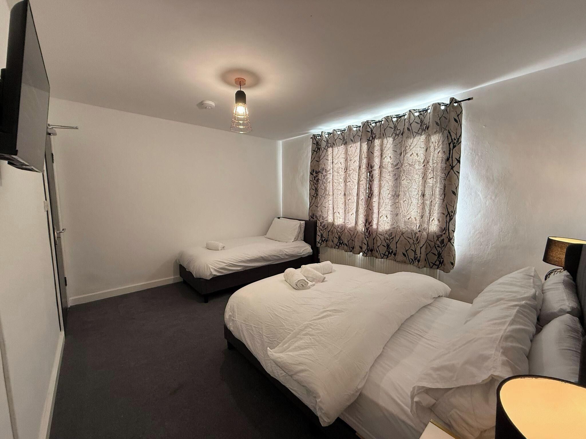 A hotel room with two single beds, a large bed, a ceiling light, and a window with patterned curtains.