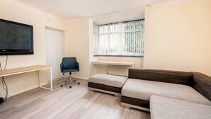 Ein modernes Wohnzimmer mit großem Flachbildfernseher, einem Holztisch, einem blauen Bürostuhl, einem braunen Sofa und einem Fenster mit Jalousien.