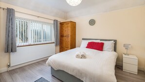 Ein Schlafzimmer mit Bett, Holzschrank, Nachttisch mit Lampe, Fenster mit Vorhängen und Heizkörper.