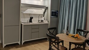 A compact kitchen with a sink, a coffee machine, and a microwave. There is a dining table with chairs and a fruit bowl. The room has wooden flooring and a blue curtain.