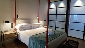 A four-poster bed with white linens and a green throw, a bedside table with a lamp, and a view through a window with frosted glass panels.
