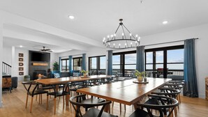 A spacious dining area with a long wooden table and black chairs, a chandelier, and large windows offering a view of the ocean.