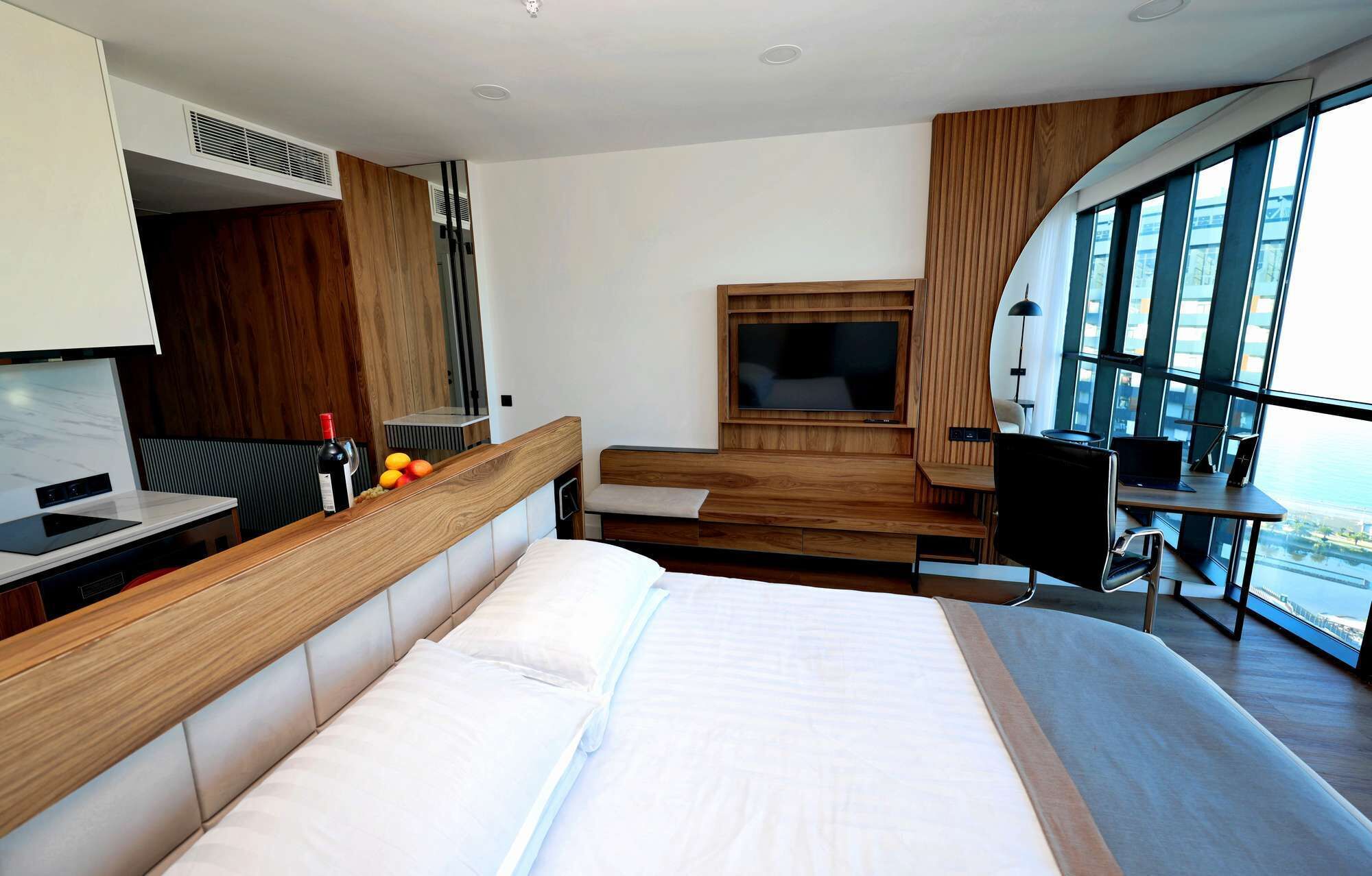 A modern hotel room with a large bed, a desk with a laptop, a TV, and a balcony with a view.