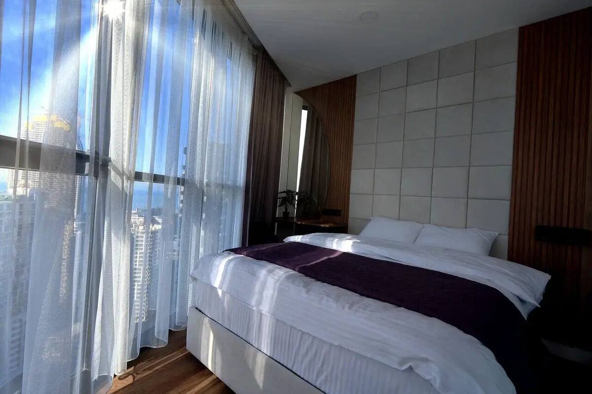 Un dormitorio con una cama grande, cabecera de madera y vistas a un paisaje urbano a través de cortinas de gasa.