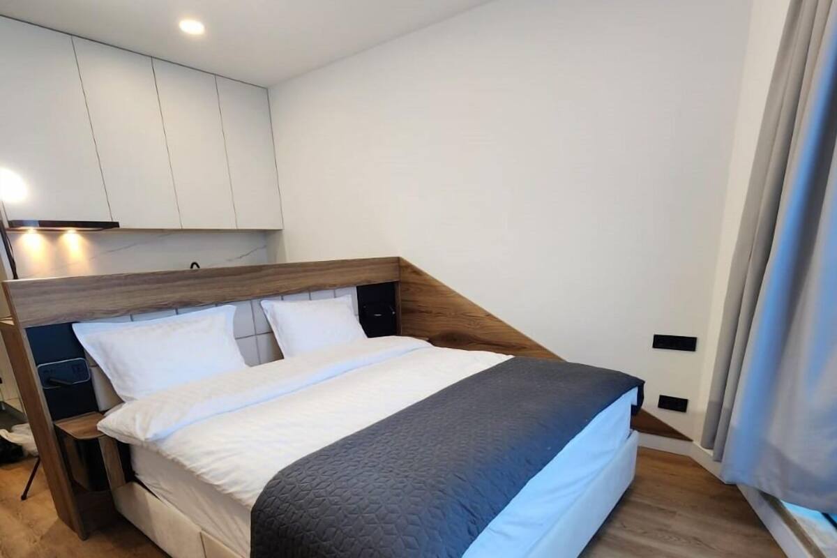 Un dormitorio moderno con cabecera de madera, una cama grande con ropa de cama blanca y oscura, una mesita de noche y un armario empotrado.