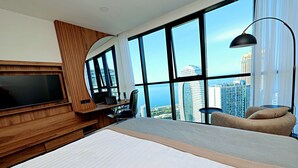 Quarto de hotel moderno com uma cama grande, uma escrivaninha com um laptop, uma televisão e vista para o horizonte da cidade.