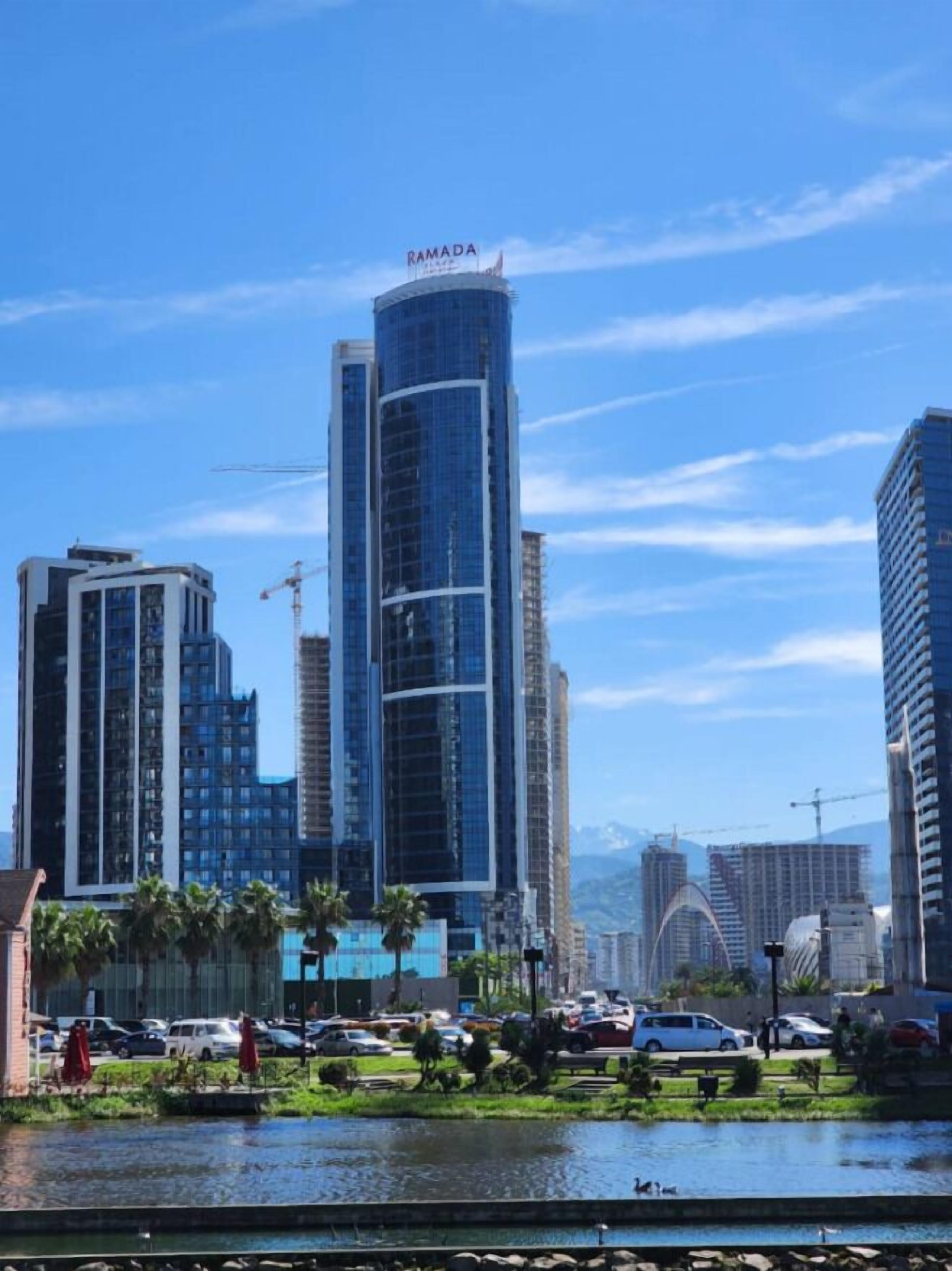 A modern high-rise building with the name 'RAMADA' on top, surrounded by other high-rise buildings and a body of water in the foreground.