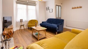 Ein Wohnzimmer mit Fernseher, einem gelben Sessel, einem blauen Sofa, einem Holztisch und einer Hängelampe.