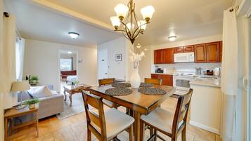 A dining area with a wooden table, chairs, a chandelier, and a kitchen in the background.