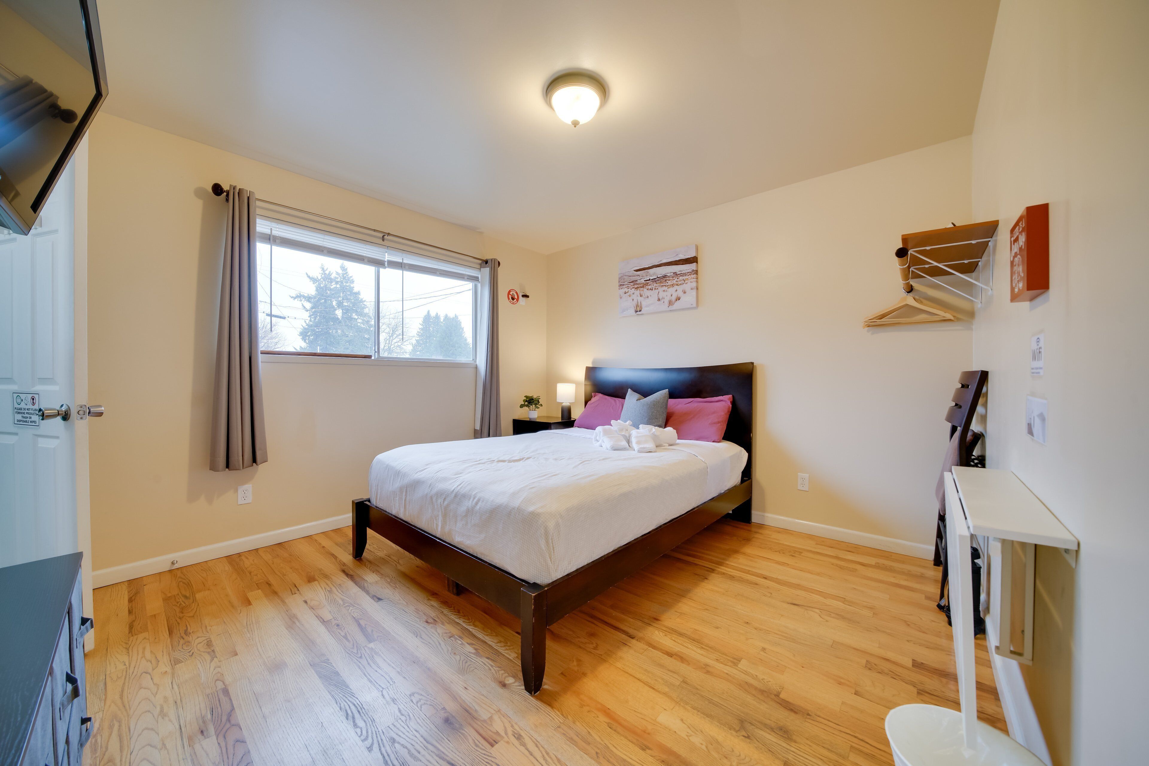 A bedroom with a wooden floor, a bed with white bedding, a nightstand, and a window with curtains.