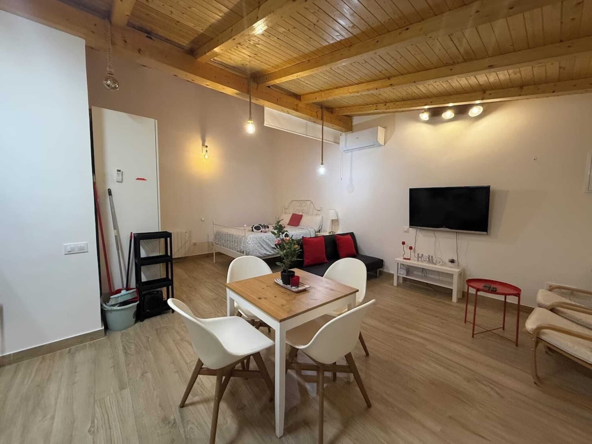 A modern, well-lit room with a wooden ceiling, a bed with red pillows, a TV, and a dining area with white chairs and a wooden table.