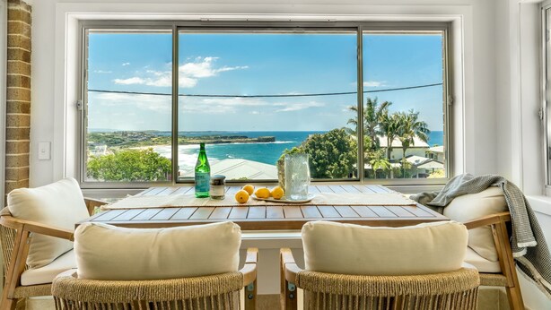 A room with a large window overlooking a beach and ocean, featuring wicker chairs, a table with a bottle and lemons, and a glass.