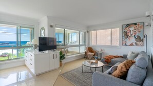 A modern living room with a large TV, comfortable seating, a view of the ocean, and a balcony with a railing.