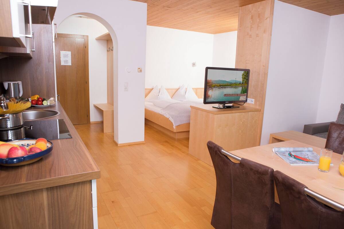 A compact room with a bed, a TV, a small kitchen area with a sink and a fruit bowl, and a wooden dining table.