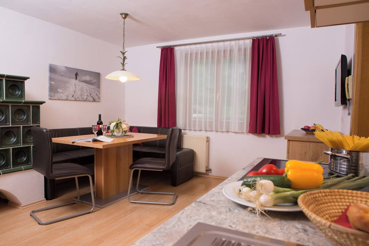 A cozy living room with a wooden dining table, black leather sofa, and a large window with sheer curtains.
