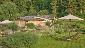 A wooden cabin with a deck, surrounded by lush greenery and a pond. - Landhaus Huber (Altenmarkt im Pongau)
