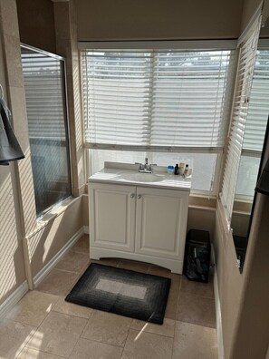 A small bathroom with a white vanity, a window with blinds, and a black doormat.