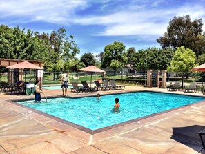 A swimming pool with lounge chairs and umbrellas, surrounded by trees and a fenced area.