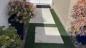 A narrow pathway leading to a white door with a glass panel, flanked by green artificial grass and potted plants. - Studio in QUIET  SFR home, gated community. (Laguna Niguel)