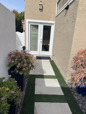 A narrow pathway leading to a white door with a glass panel, flanked by potted plants and a small table.