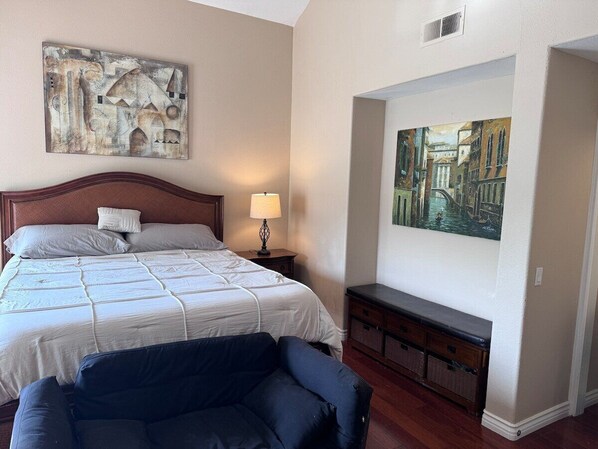 A bedroom with a large bed, a nightstand, a lamp, a bench, and two paintings on the wall.
