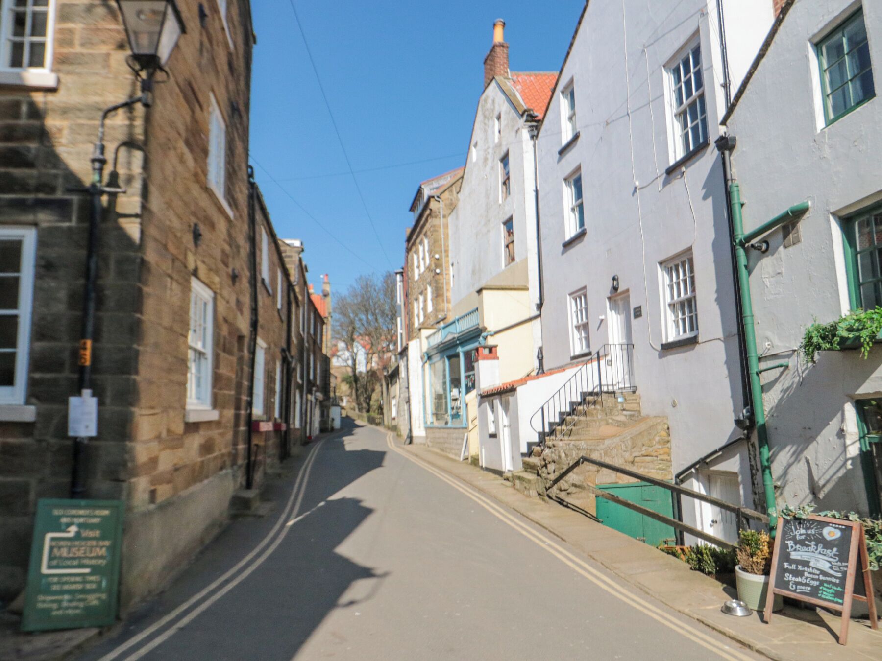 A narrow street lined with historic buildings, a sign for a museum, and a chalkboard sign advertising breakfast.