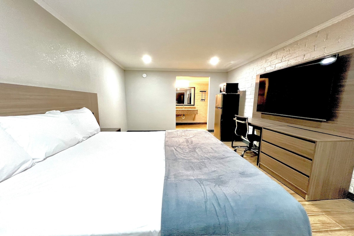 A hotel room with a large bed, a desk with a chair, a flat-screen TV mounted on the wall, and a kitchenette area in the background.