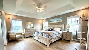 A bedroom with a large bed, a dresser, a chair, a nightstand, a ceiling fan, and a window with curtains.