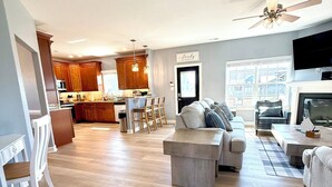 A spacious living area with a large sofa, a coffee table, and a dining area with chairs. There is a kitchen in the background with wooden cabinets and a ceiling fan.