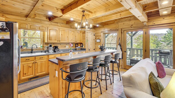 A wooden cabin with a kitchen featuring a bar area, wooden cabinets, and a large window overlooking a deck. - 3 Mi to Dtwn Clayton: Cabin w/ Trail Access! (Clayton)