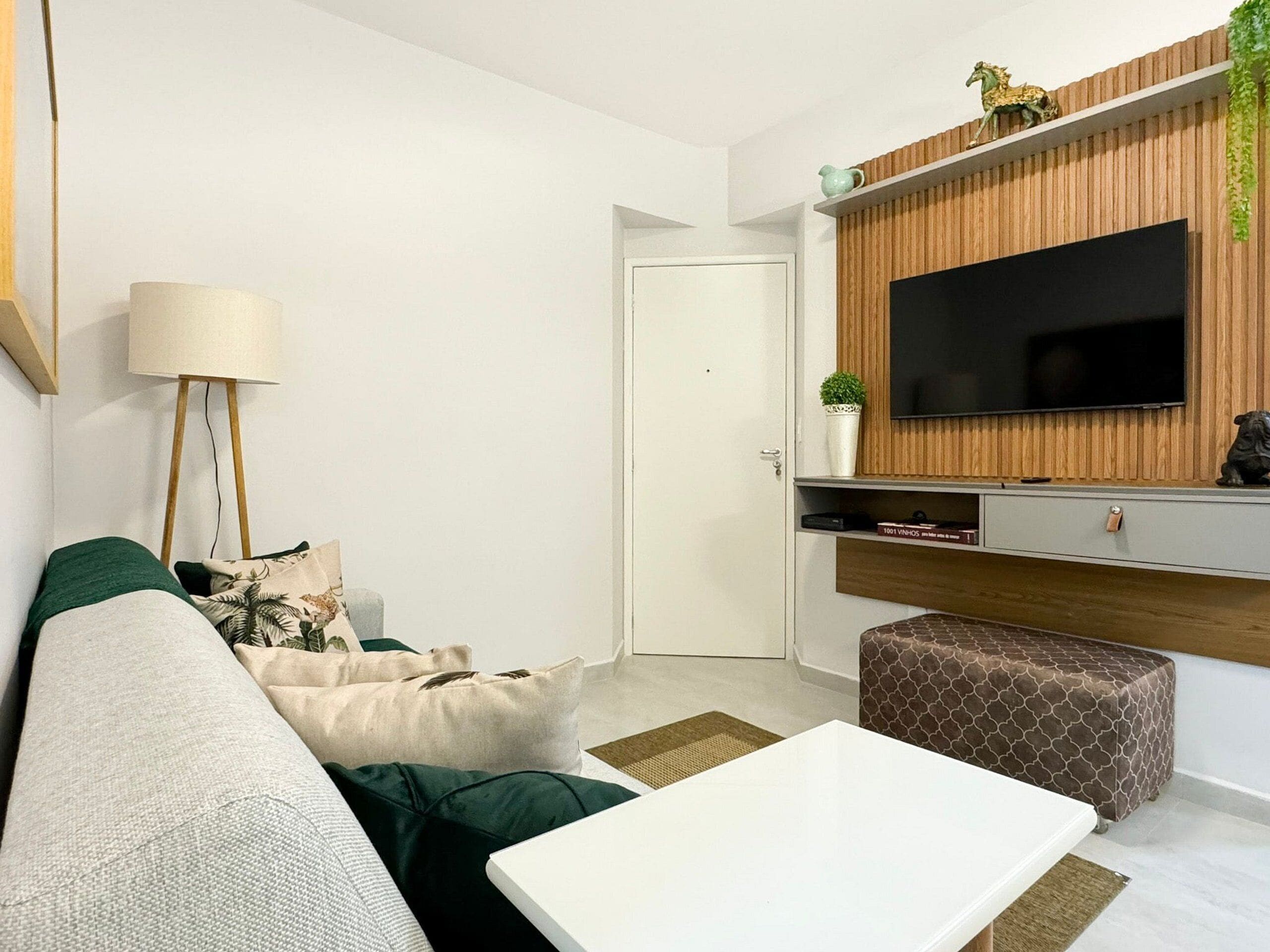 A modern living room with a white sofa, a coffee table, a TV mounted on a wooden panel wall, a lamp, and a small plant.