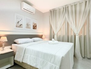 A hotel room with a neatly made bed, two bedside lamps, a wall-mounted air conditioner, and a set of curtains.