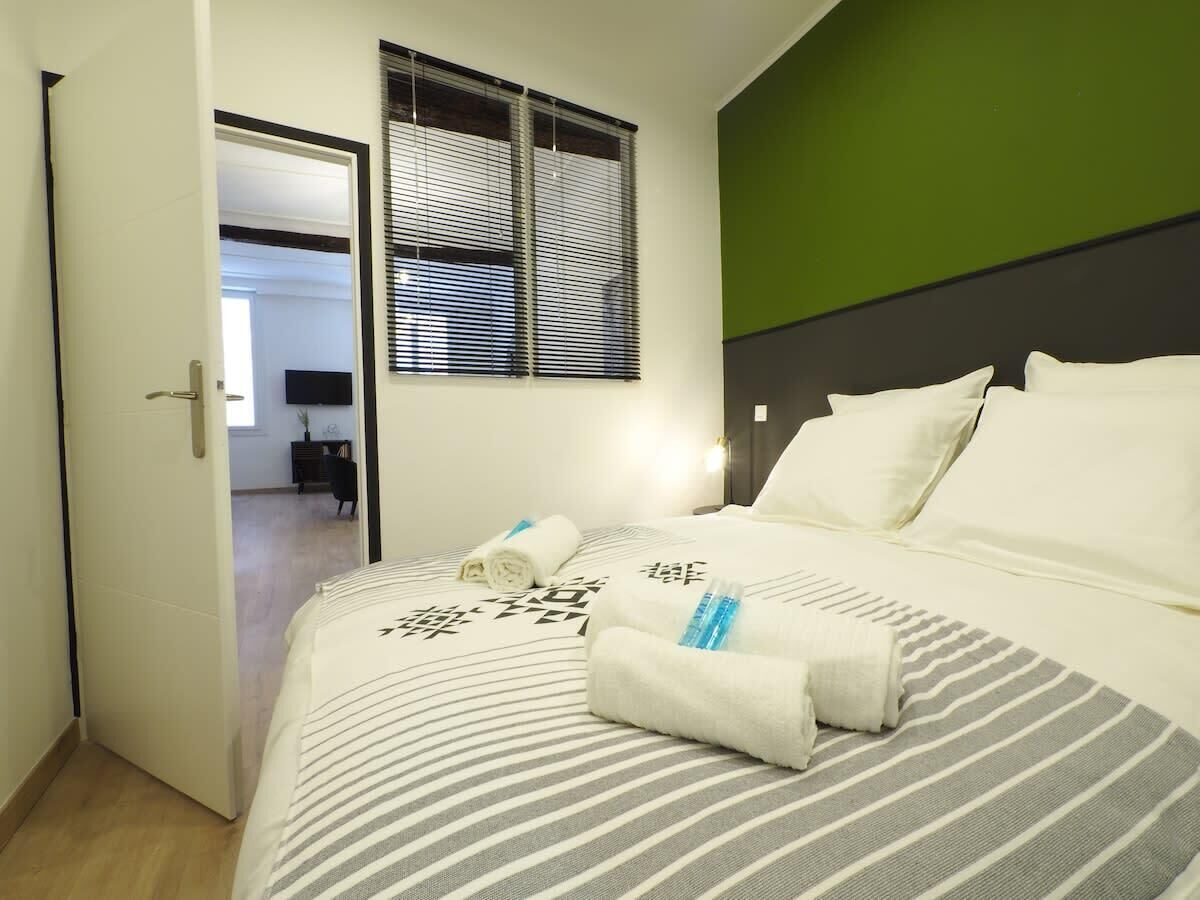 A hotel room with a bed, a green accent wall, and a window with blinds.