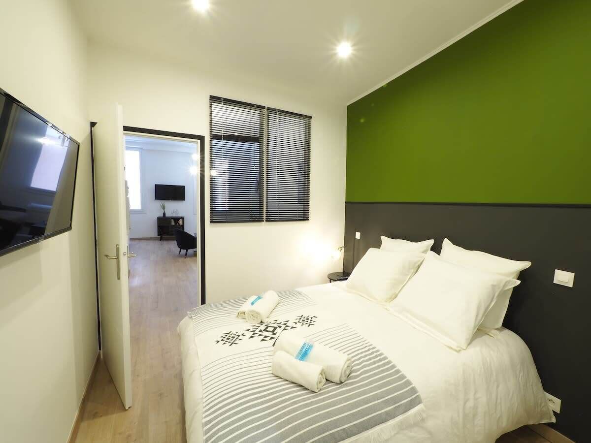 A bedroom with a bed, a TV, a window with blinds, and a green accent wall.