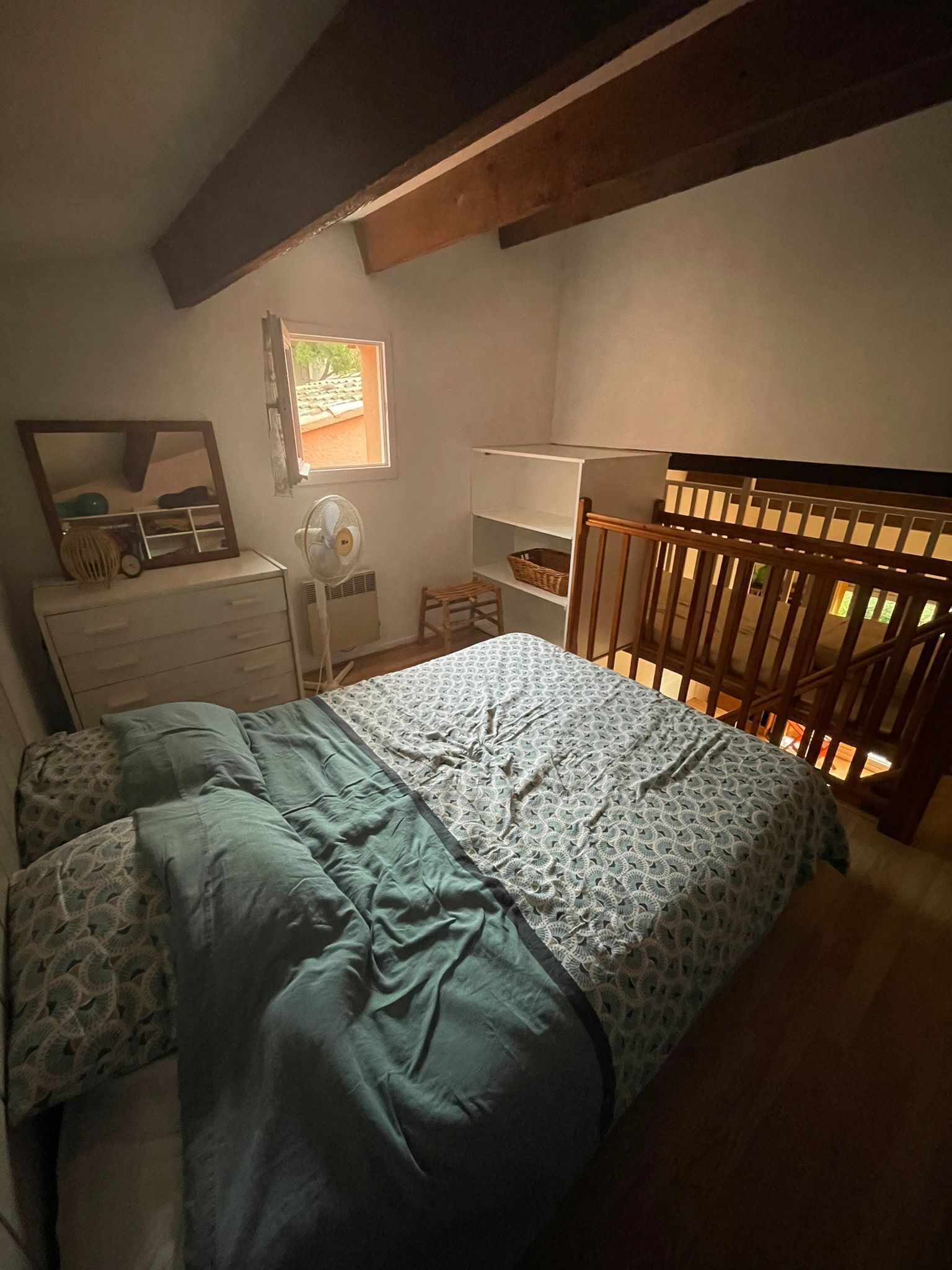 A bedroom with a bed, dresser, mirror, fan, and a window with a view of greenery.