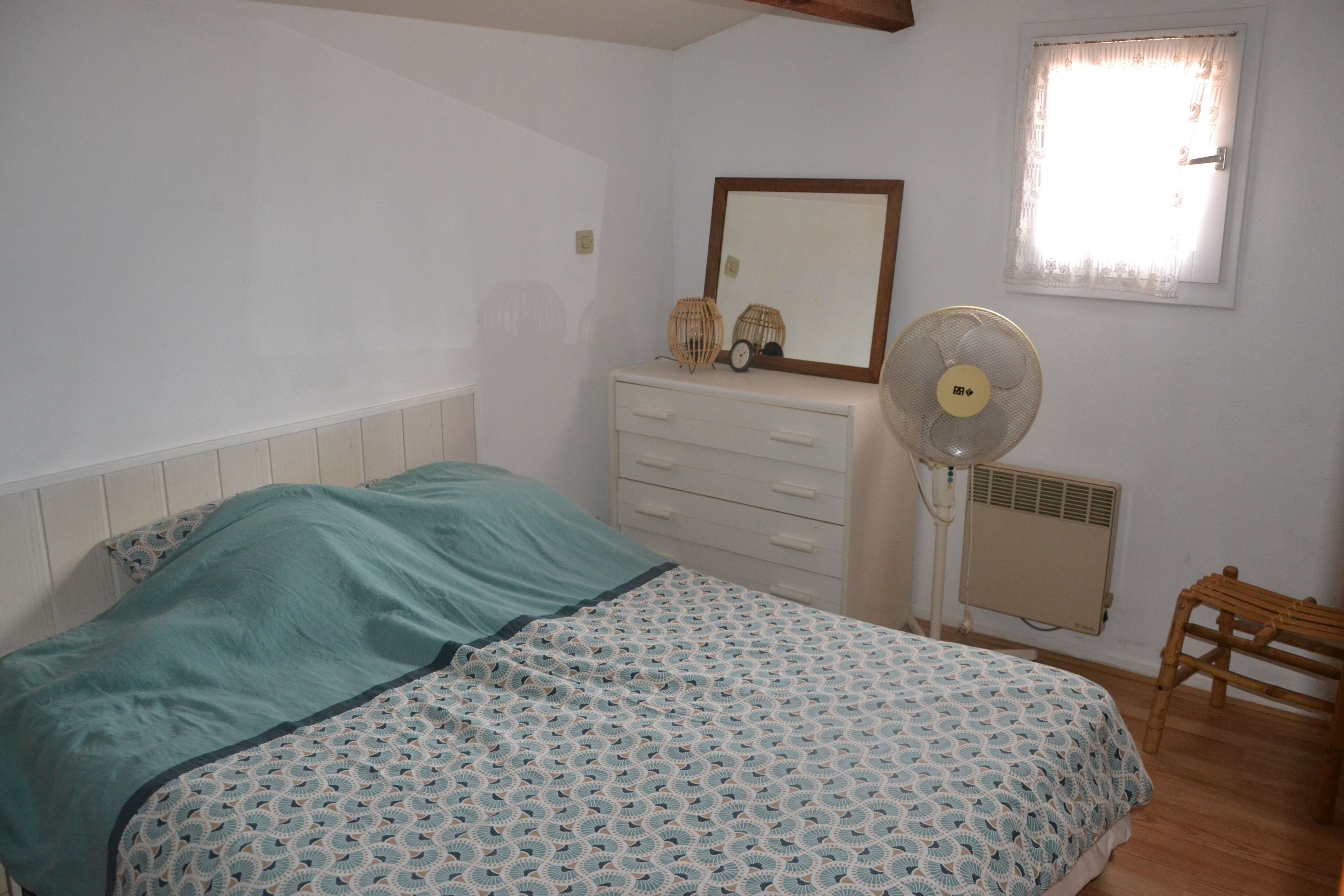 A bedroom with a bed, a dresser, a fan, a window, and a radiator.